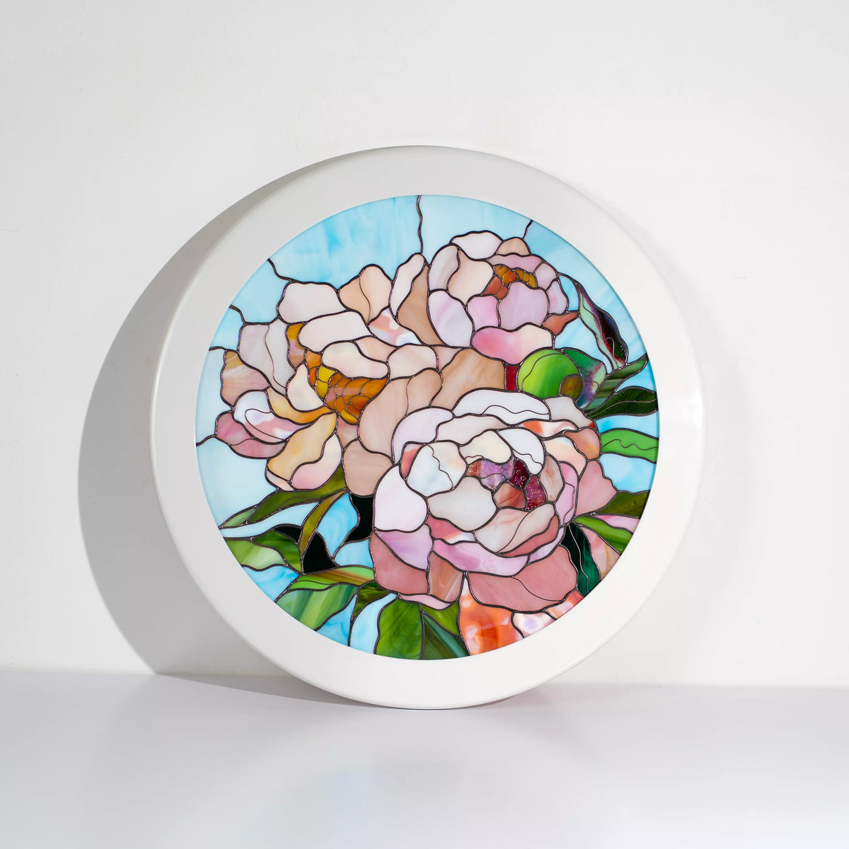 Round framed stained glass peony panel leaning against a white wall, pink and cream peonies on a soft blue sky background - handmade floral stained glass wall art and thoughtful gift for mom
