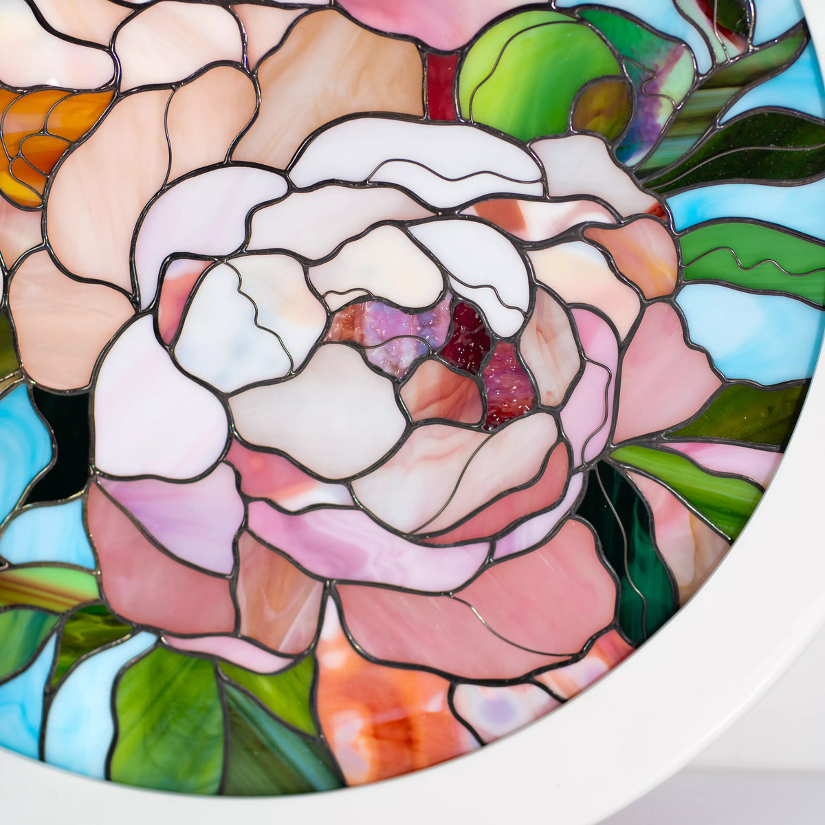 Close-up of a handmade stained glass peony panel showing blush and peach petals, amber stamens and dark soldered copper foil seams - round framed floral glass wall art in a white frame