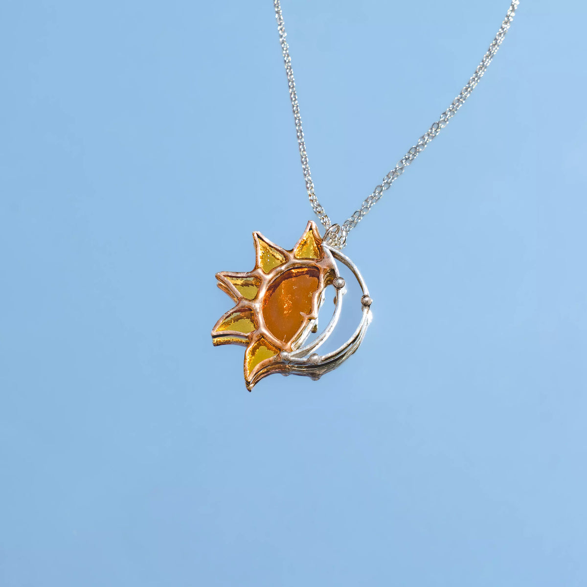 Handmade stained glass sun and moon necklace against a blue sky - a glowing amber sun with pointed rays cradled by a silver crescent moon, on a fine stainless steel chain