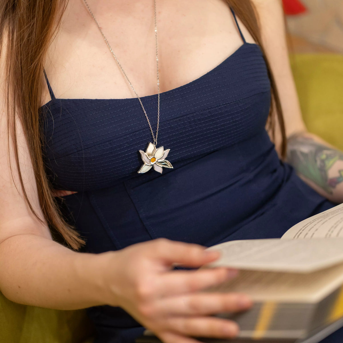 White Lotus stained glass necklace worn with a navy dress as a woman reads a book, the white lotus pendant resting on her chest - handmade floral stained glass jewelry and a serene gift