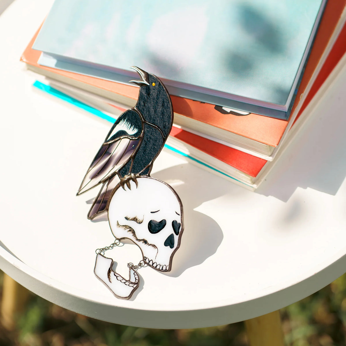 Handmade stained glass raven and skull suncatcher on a sunlit table beside a stack of books, a cawing black raven with an iridescent lilac wing on a white skull with a hinged jaw on chains - gothic Halloween decor