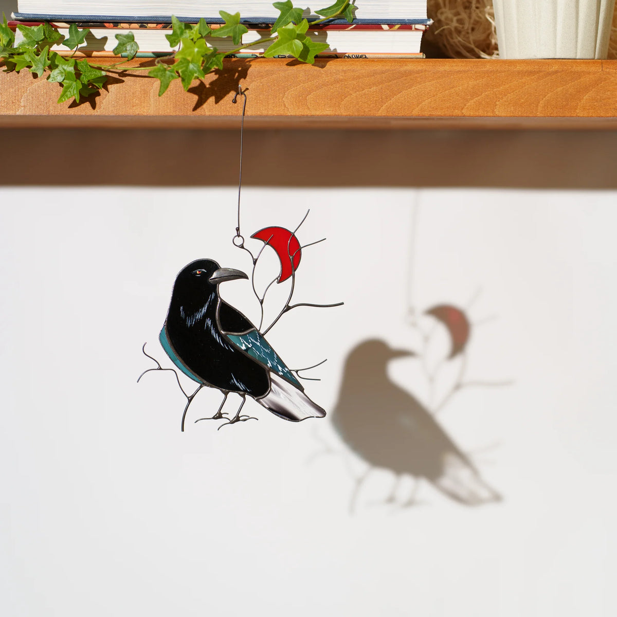 Stained glass raven with a red crescent moon suncatcher hanging by a shelf of books and ivy, casting a crisp shadow in sunlight - black raven with an iridescent teal wing on wire branches, gothic window decor