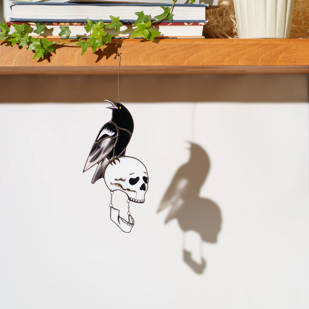 Stained glass raven and skull suncatcher hanging below a shelf of books and ivy, casting a crisp shadow on a white wall - cawing black raven on a bone-white skull with a swinging jaw, gothic Halloween window decor