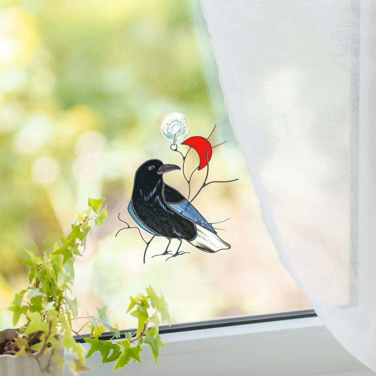 Raven with red crescent moon stained glass suncatcher on a sunlit window with a suction hook, ivy below and a green garden beyond - black raven with an iridescent teal wing, handmade gothic window decor
