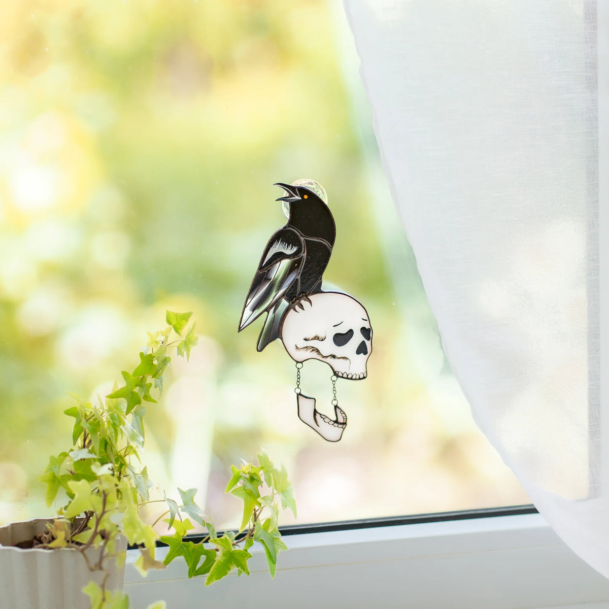 Stained glass raven-and-skull suncatcher in a bright window with sheer curtains and greenery, glossy black raven with an iridescent lilac wing over a white skull with a chained jaw - handmade gothic Halloween art