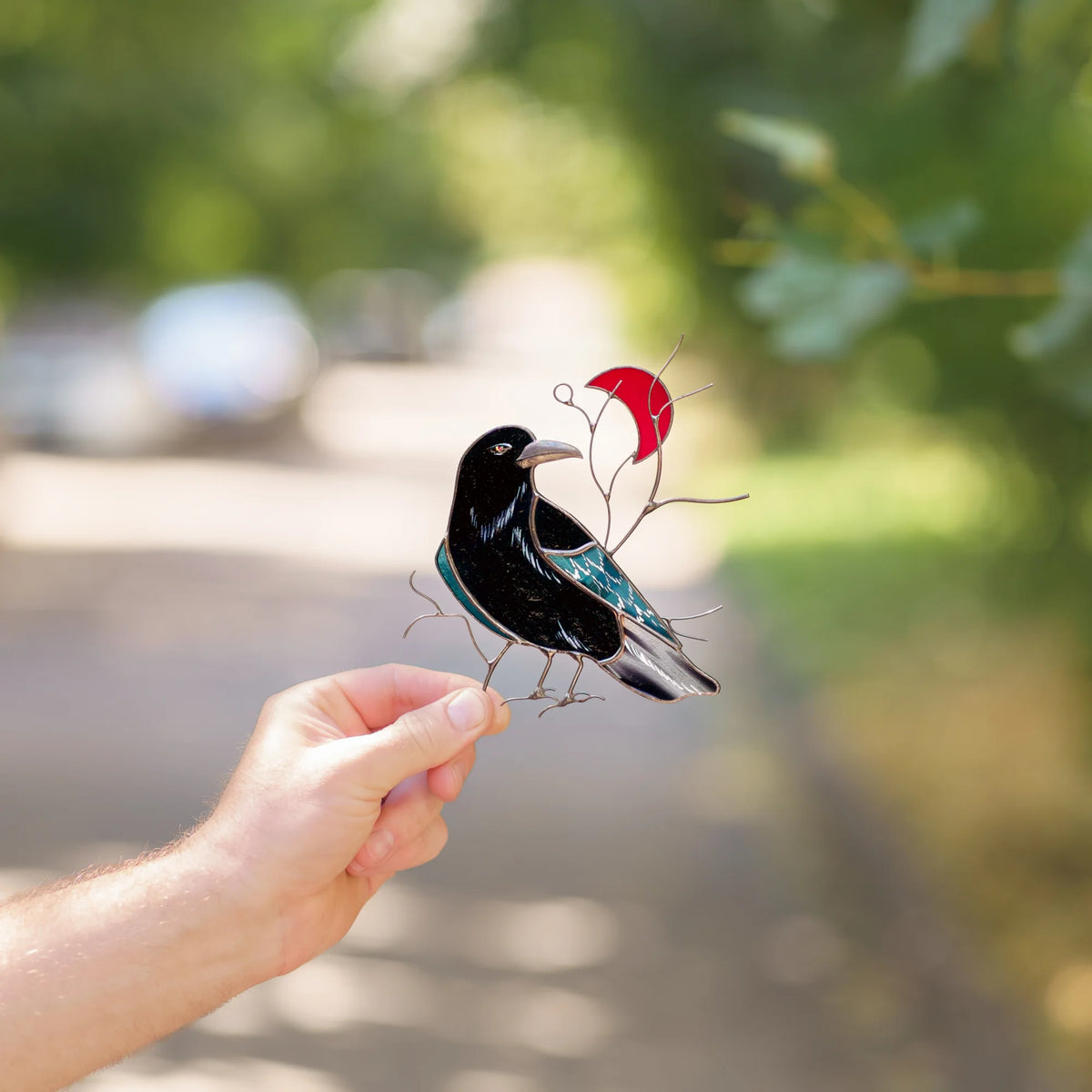 Hand holding a stained glass raven and red crescent moon suncatcher outdoors, glossy black raven with an iridescent teal wing on wire branches - handmade gothic and celestial gift, 5.5 by 5.5 inches