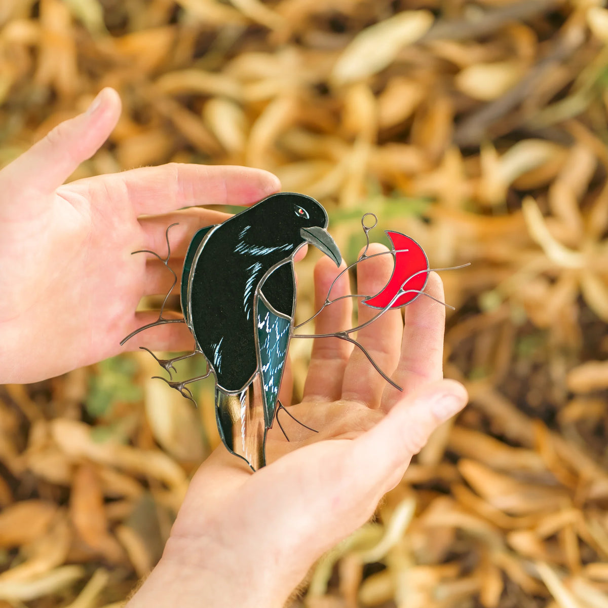 Stained glass raven with a red crescent moon cupped in two hands over autumn leaves, black raven with a teal iridescent wing and wire branches - handmade gothic suncatcher and thoughtful fall gift