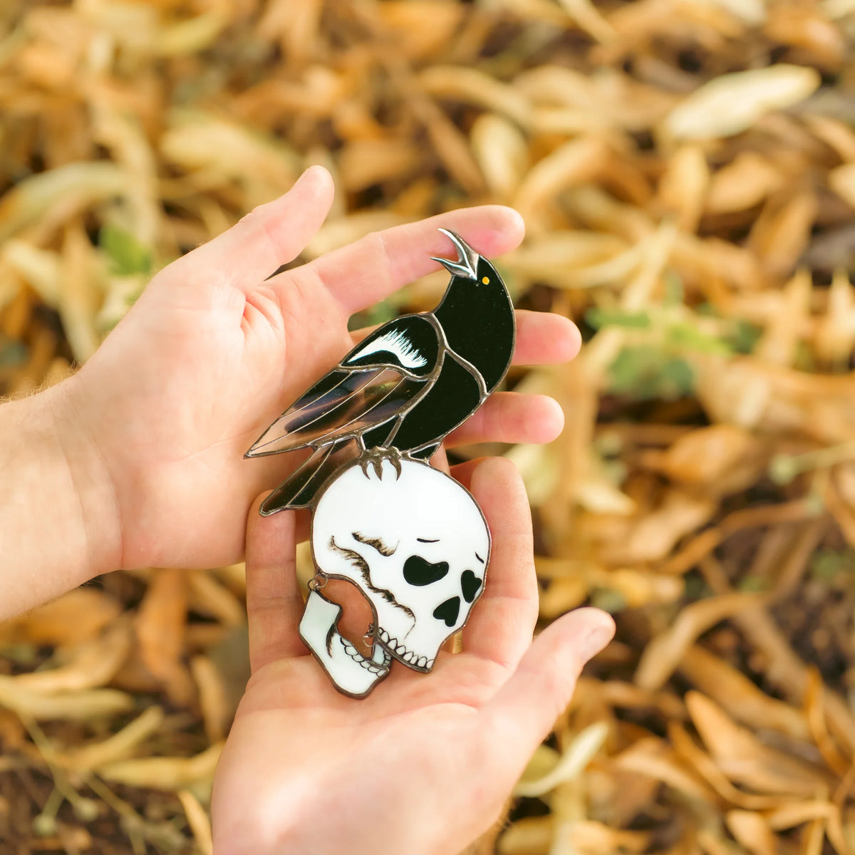 Stained glass raven and skull suncatcher held over autumn leaves, black raven with an iridescent lilac wing perched on a bone-white skull with a swinging jaw - handmade gothic memento-mori suncatcher and fall gift