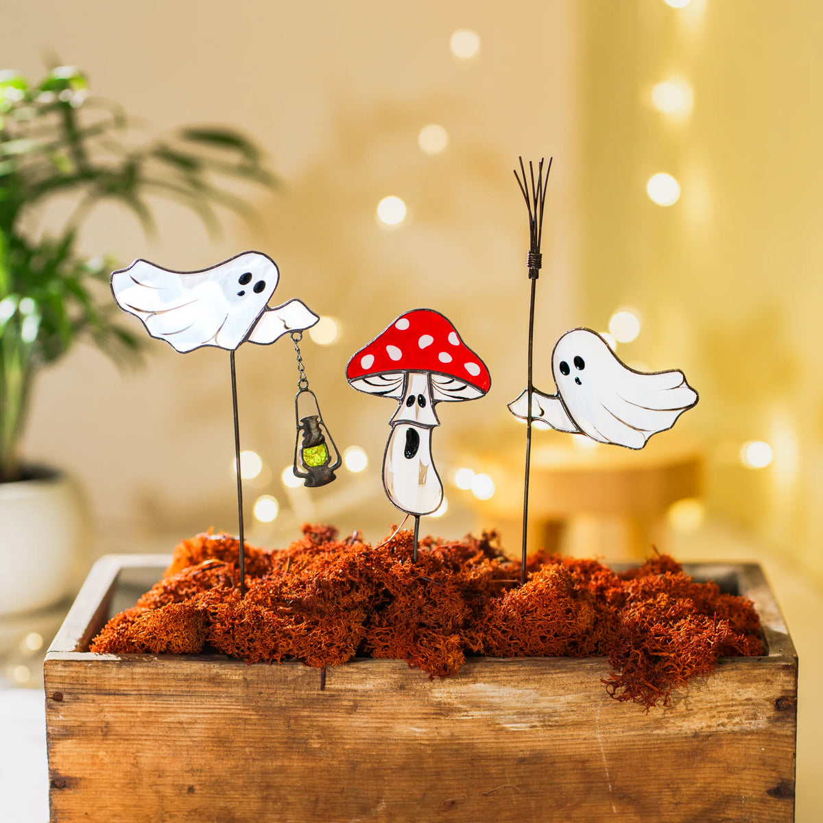 Set of three glass ghost and mushroom plant stakes in a wooden box of rust-orange moss, warm bokeh lights behind - moody stained glass halloween decor for a cozy autumn corner