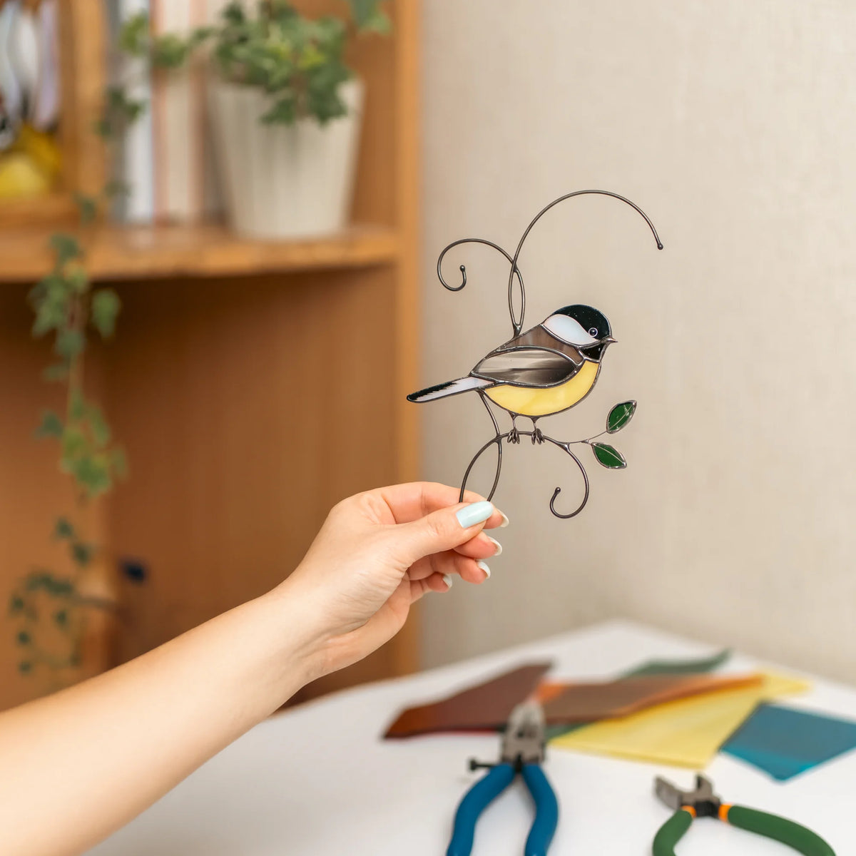 Hand holding a stained glass chickadee suncatcher in a sunlit studio with a plant shelf and glass-cutting tools - handmade black-capped chickadee with a yellow belly on a wire branch