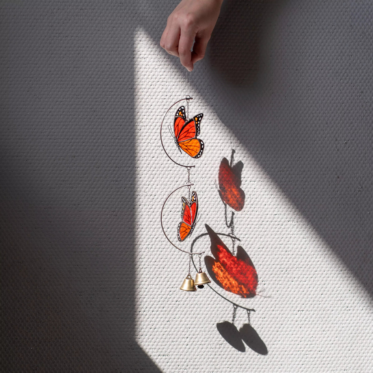 A hand holds stained glass butterfly wind chime hanging against a bright, blurred green background, highlighting the vibrant orange stained glass.
