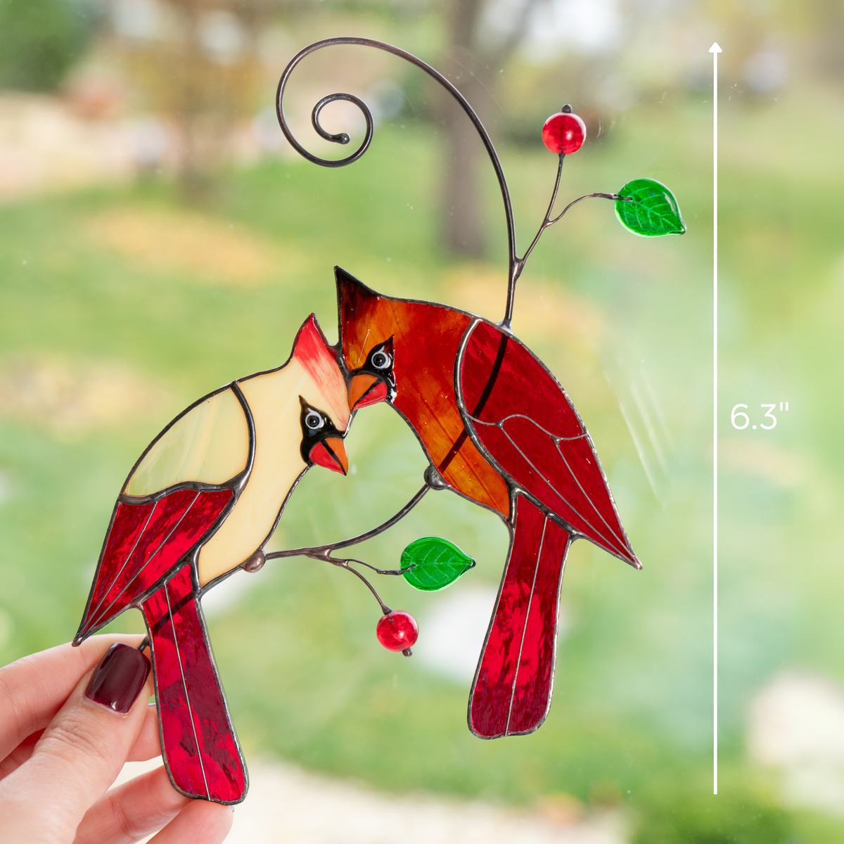 Hand holding a stained glass cardinal pair suncatcher near a window with size scale at 6.3 inches.