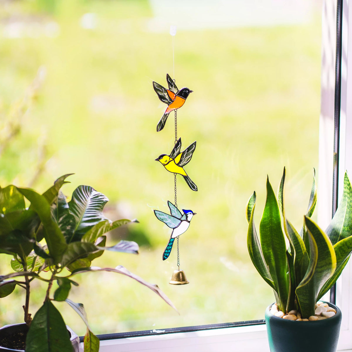 Decorative glass birds wind chime on a string hanging by a window with plants.