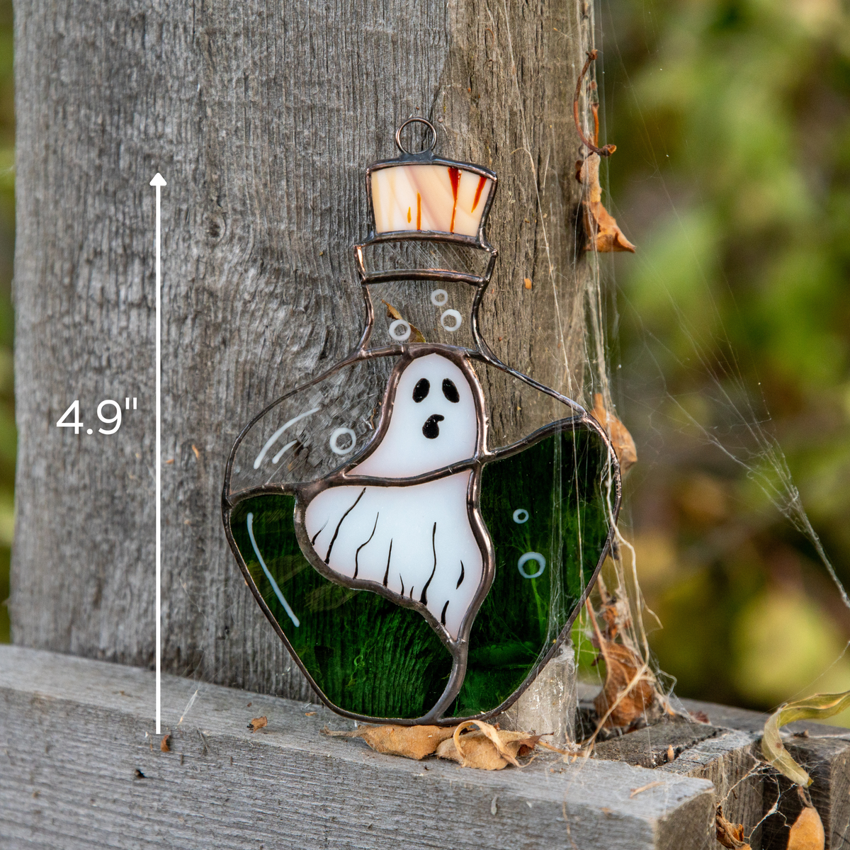 Stained glass ghost inside green potion bottle leaning against wood post outdoors with spider webs and size marking 4.9 inches.