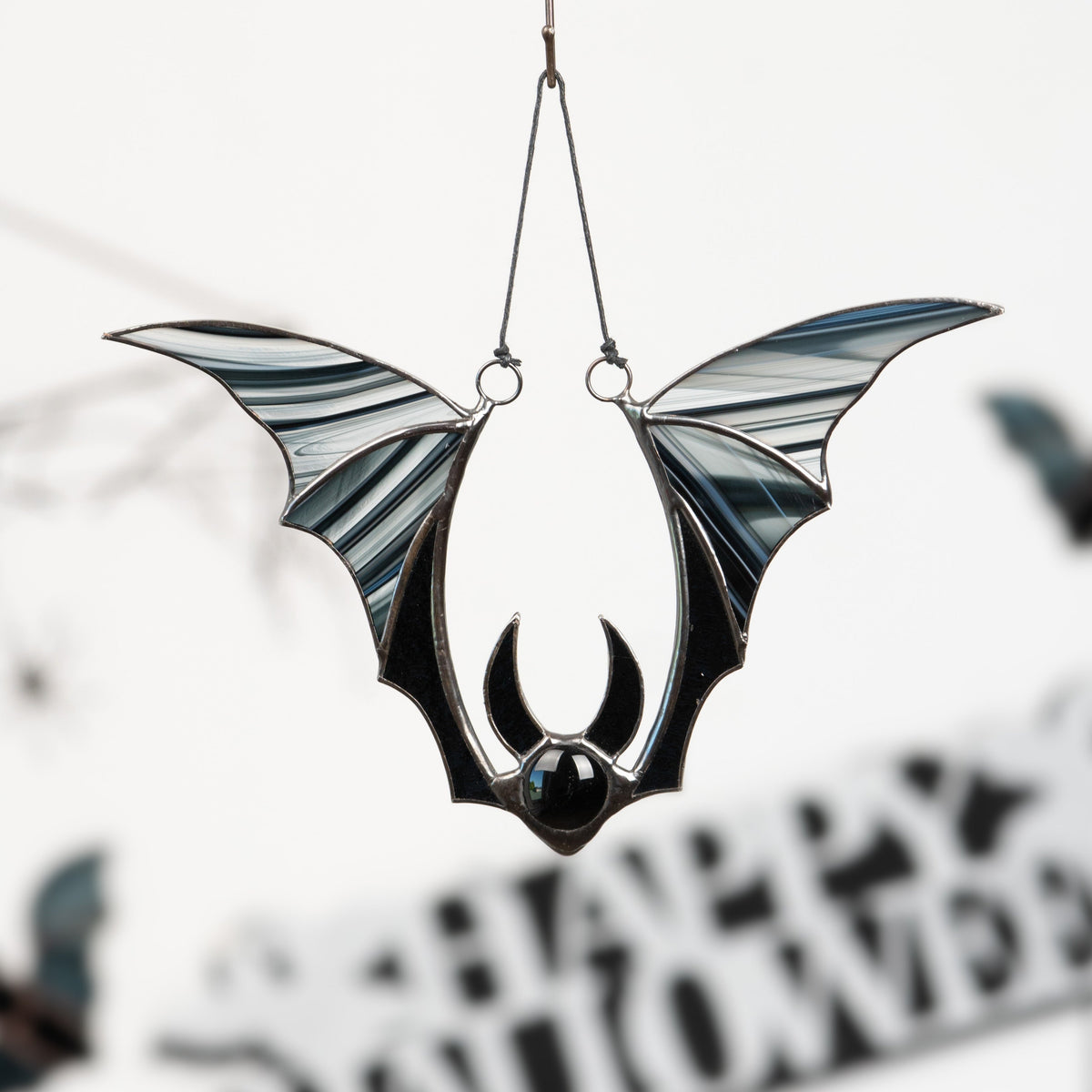 Stained glass bat with striped black wings hanging in front of a blurred "Happy Halloween" sign and Halloween-themed decor.