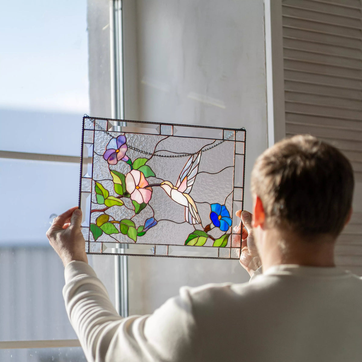 Person holding a stained glass panel with floral and bird design by a window