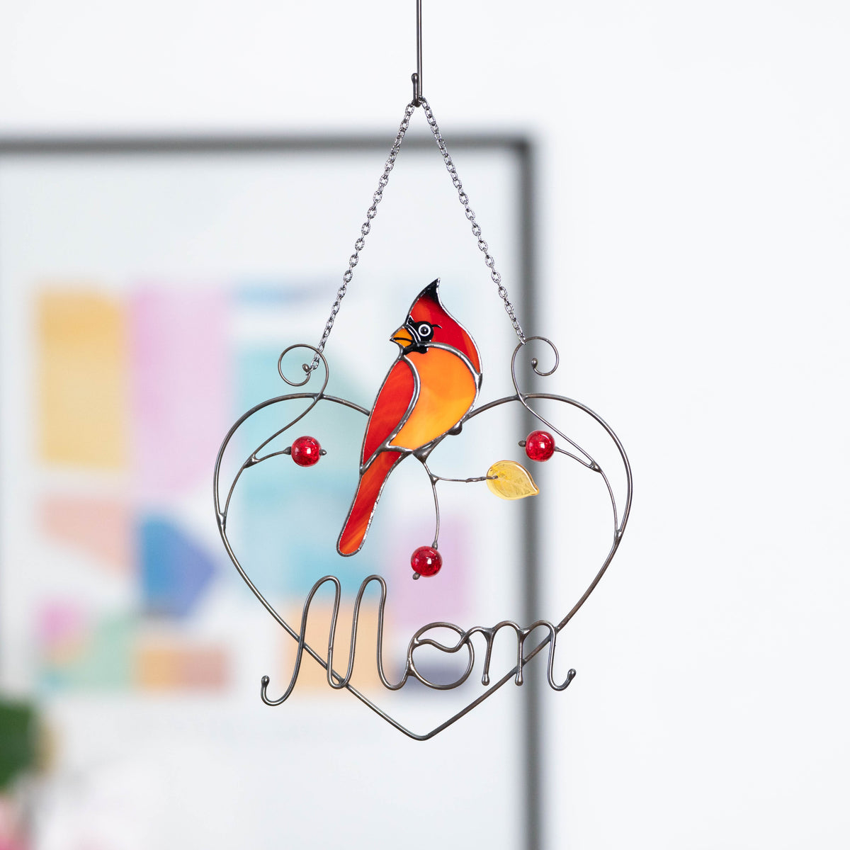 Cardinal bird suncatcher with “Mom” inscription, hanging indoors with blurred wall art behind.