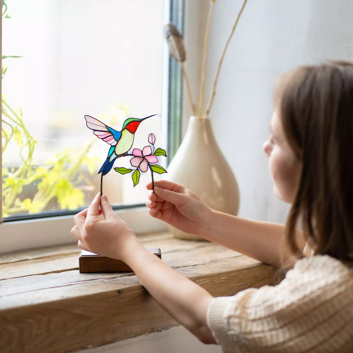 Women holding a stained glass hummingbird and flower suncatcher by a window