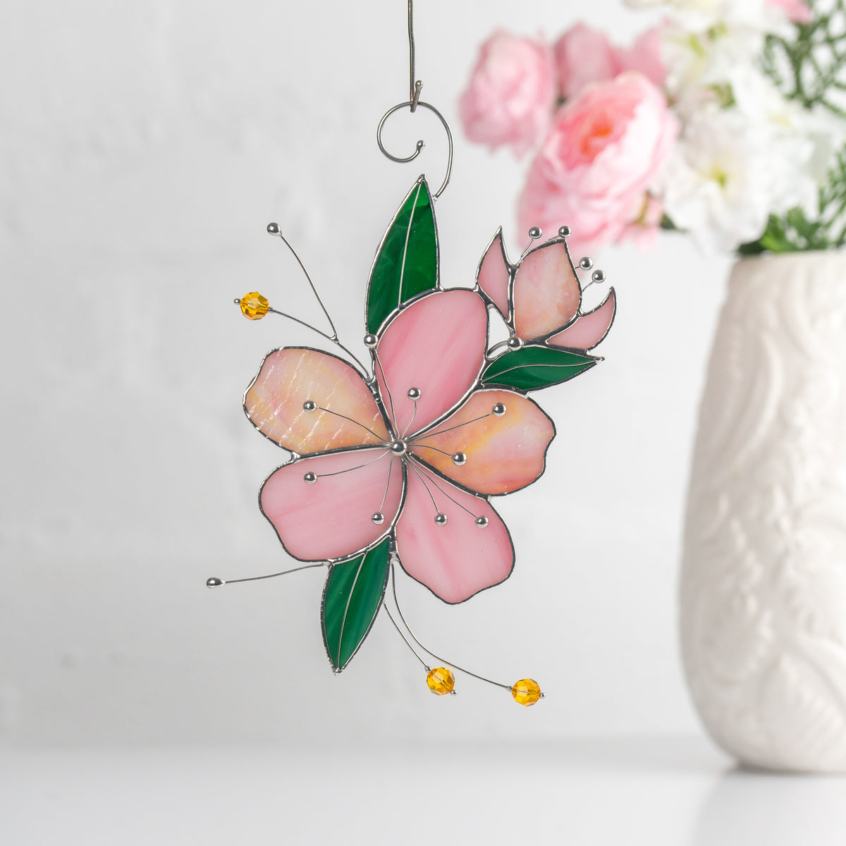 Pink stained glass sakura hanging indoors with flowers in the background, emphasizing its soft colors and artistic style.