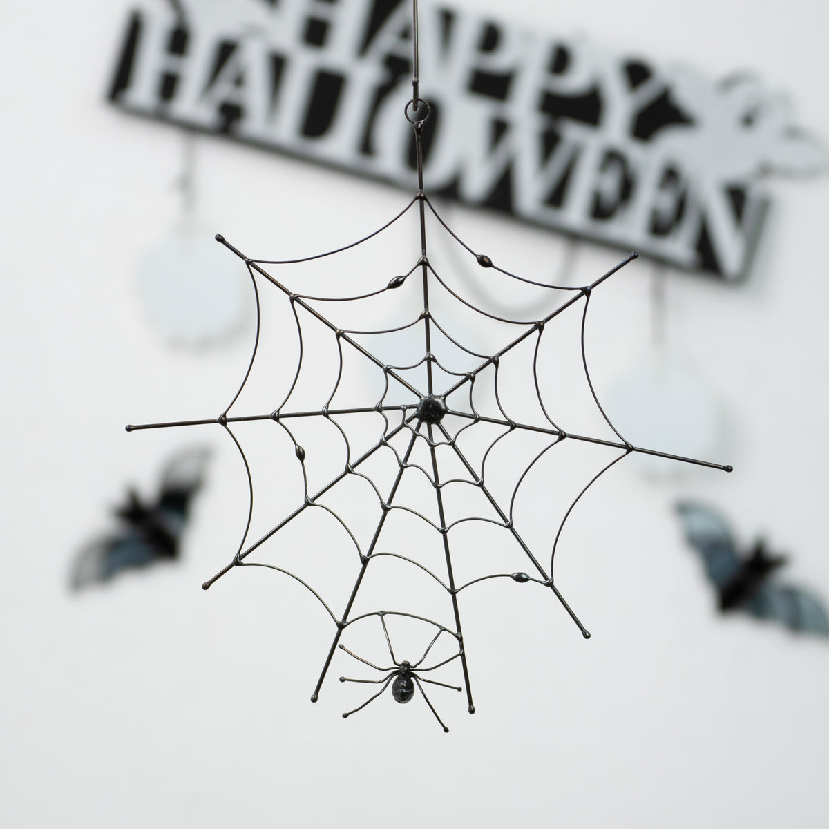 Metal spider web with a hanging spider, displayed in front of Halloween-themed wall decor with bats and a “Happy Halloween” sign.