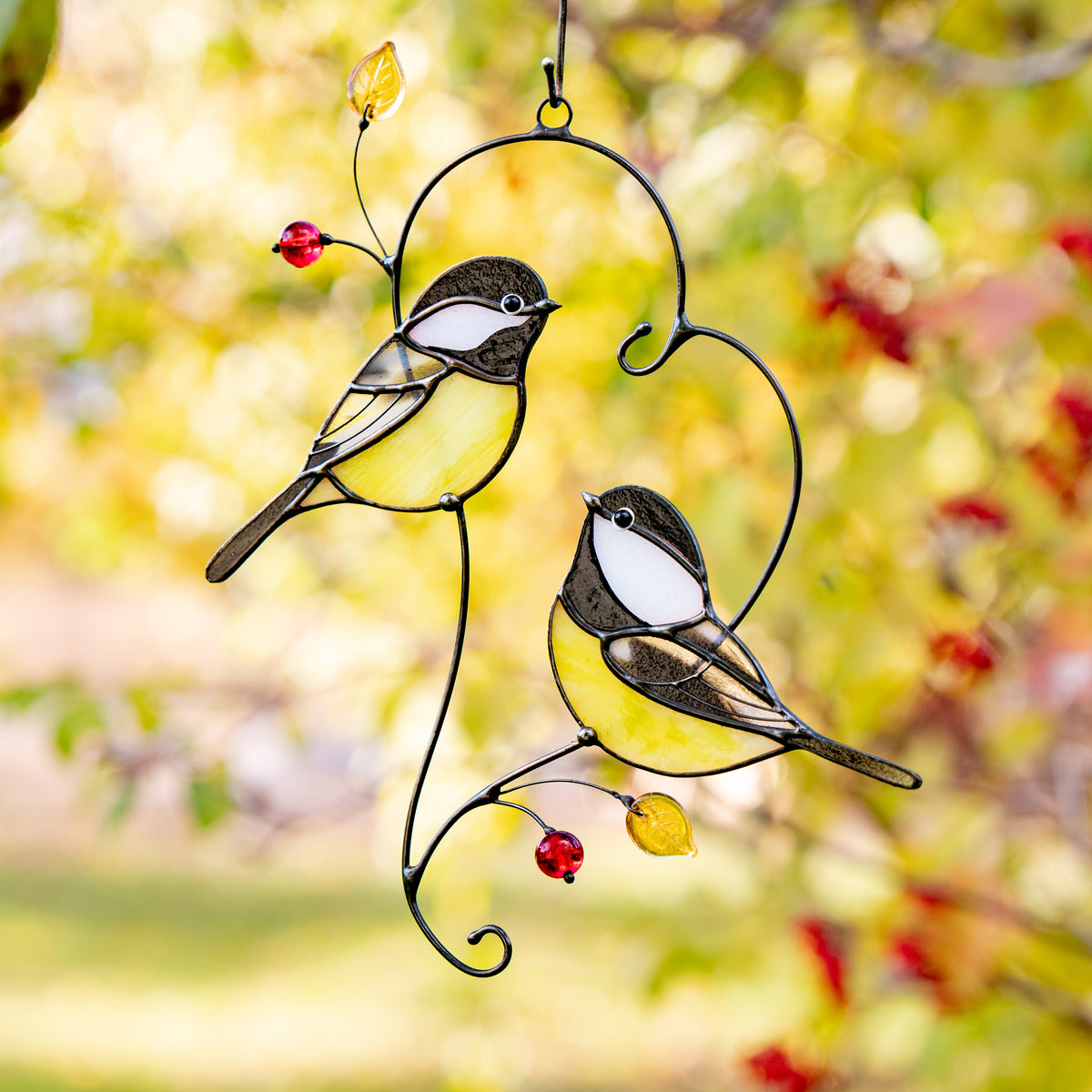 Two stained glass chickadees with yellow bellies and red beads, hanging among autumn leaves in soft golden light