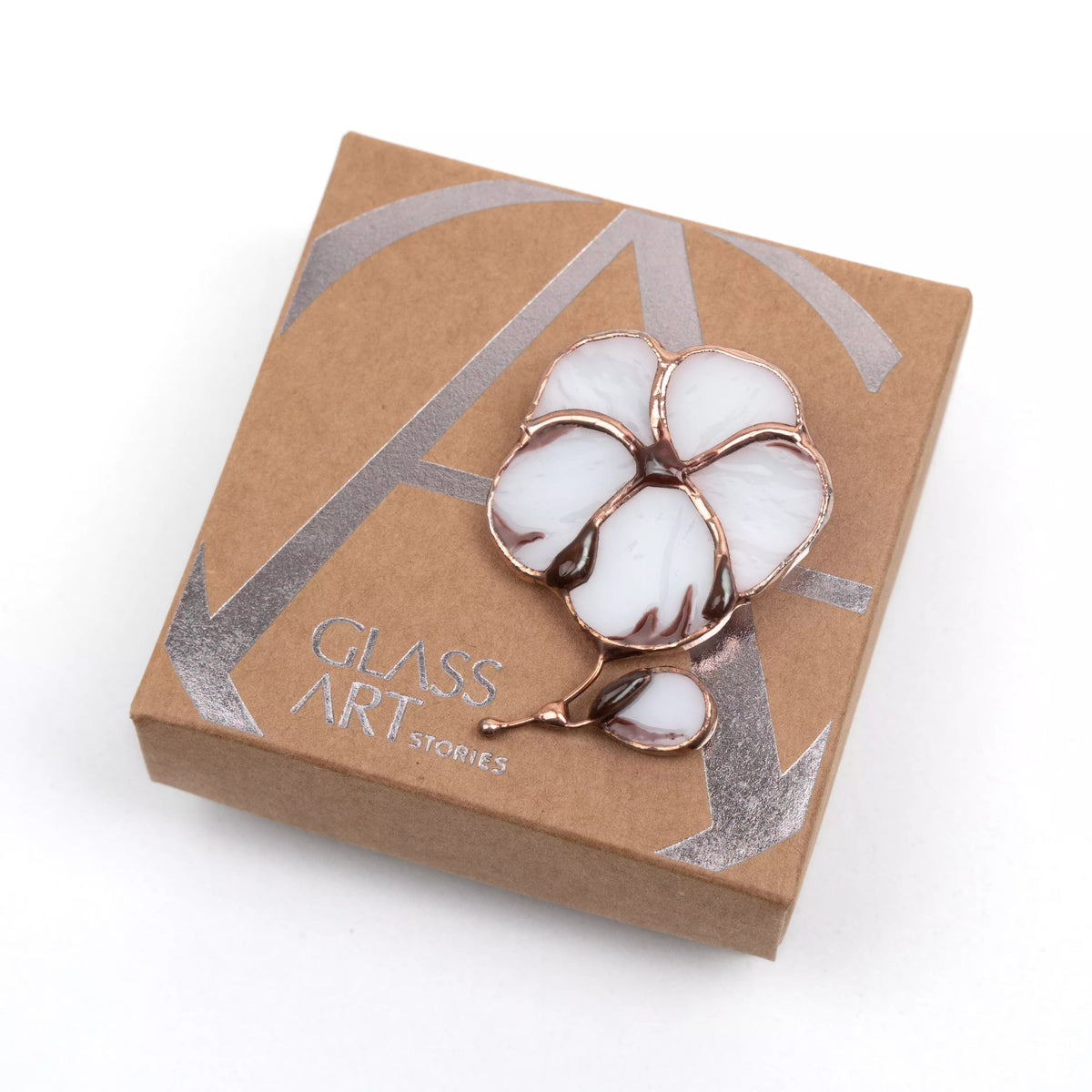 White and brown stained glass cotton brooch with copper edges, lying on a kraft gift box with silver logo.