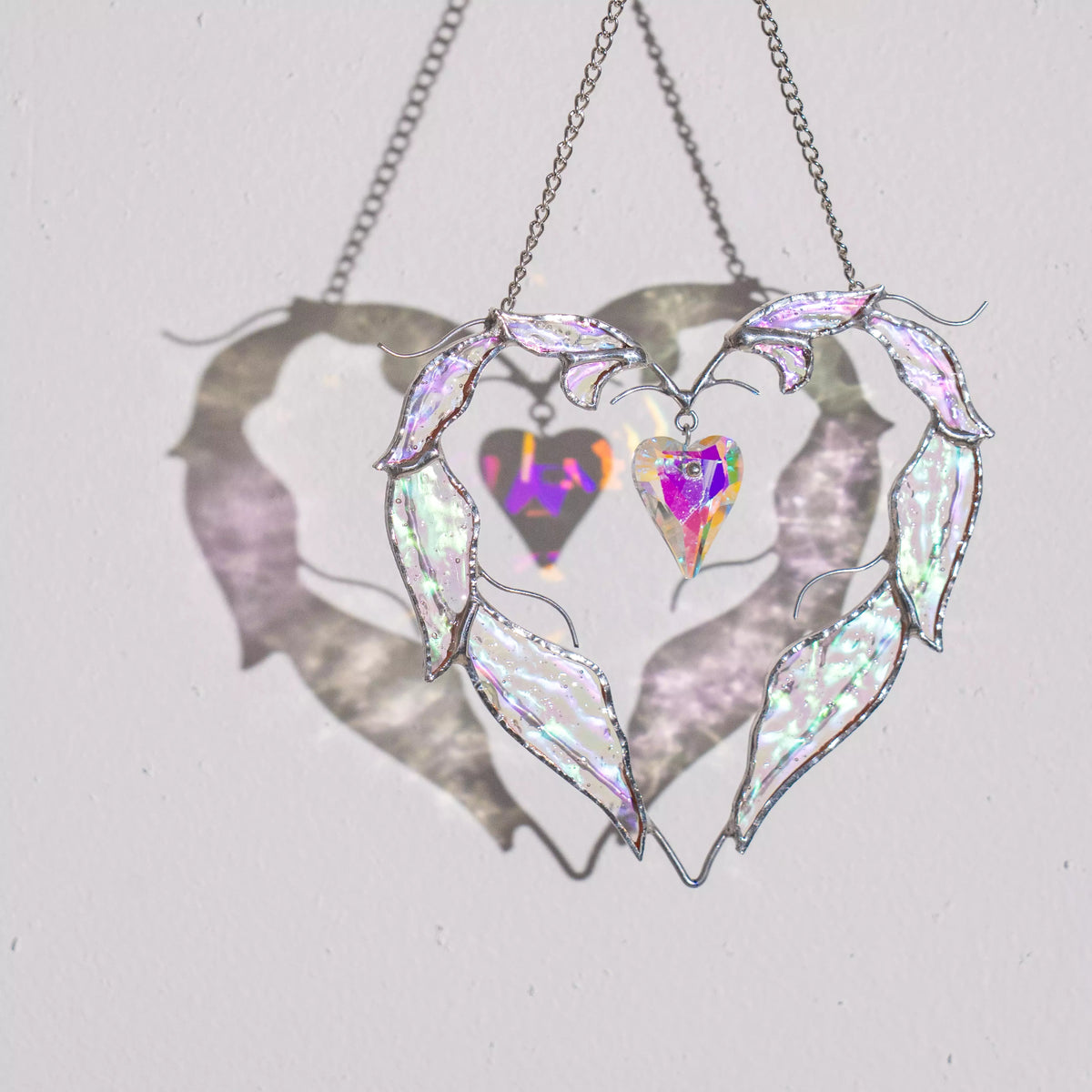 Love stained glass heart suncatcher hanging in sunlight, casting soft shadows and rainbow reflections on the wall.