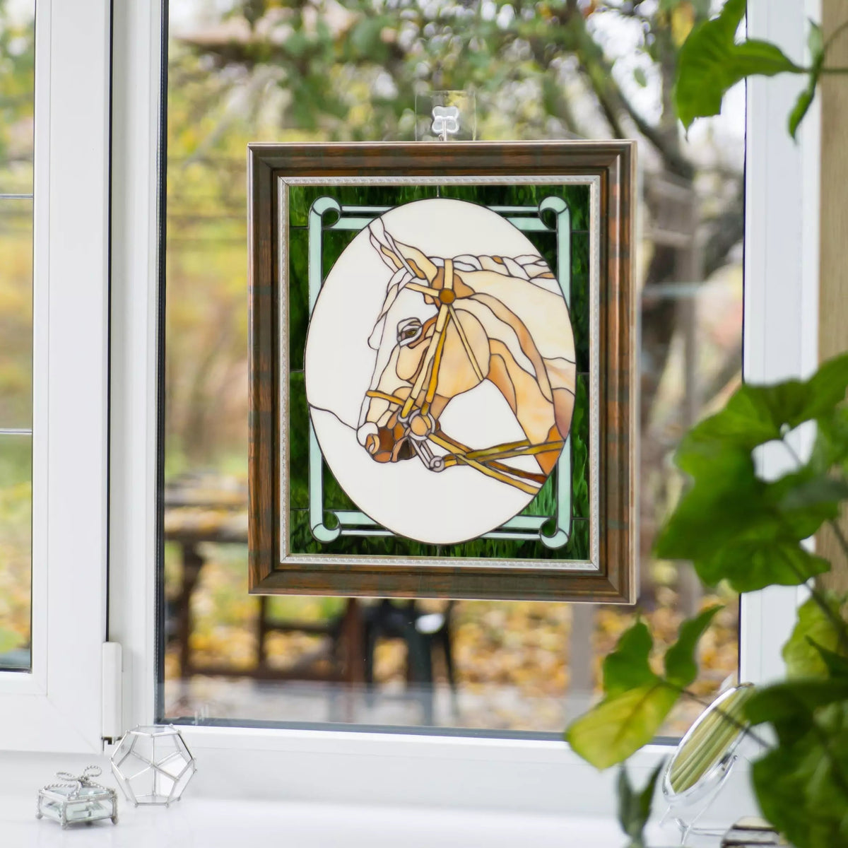 Horse stained glass panel suspended on window with sunlight shining through and greenery outside.