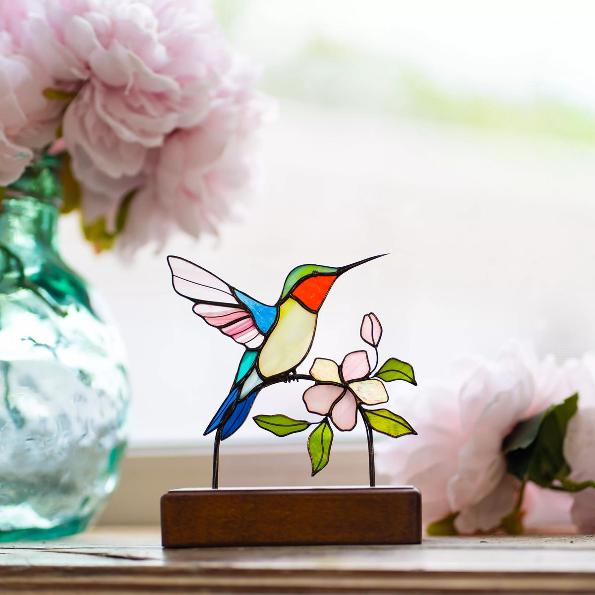 Stained glass hummingbird with flower on a wooden stand with flowers in the background