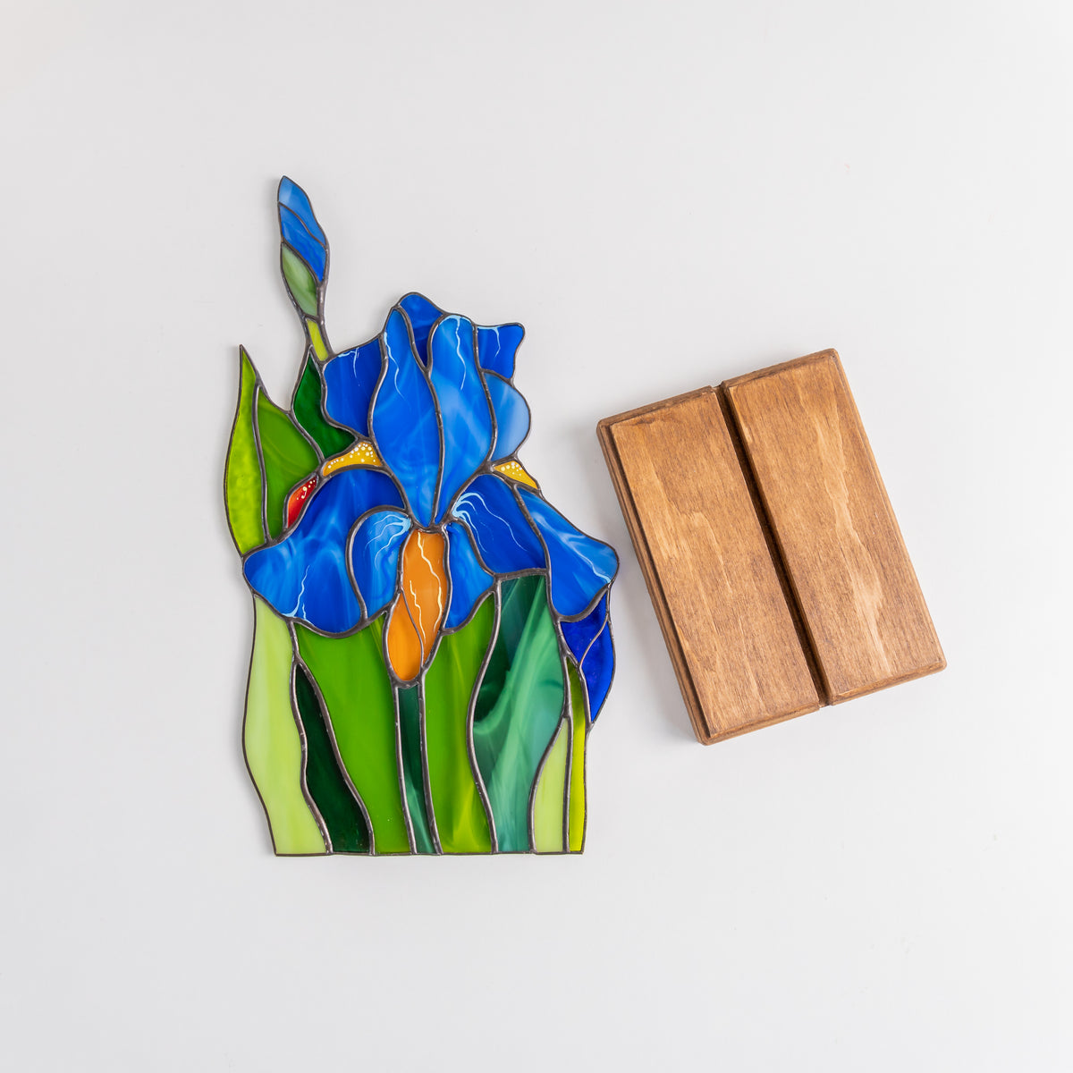 Iris flower stained glass panel laid flat beside two-piece wooden display stand on white surface.