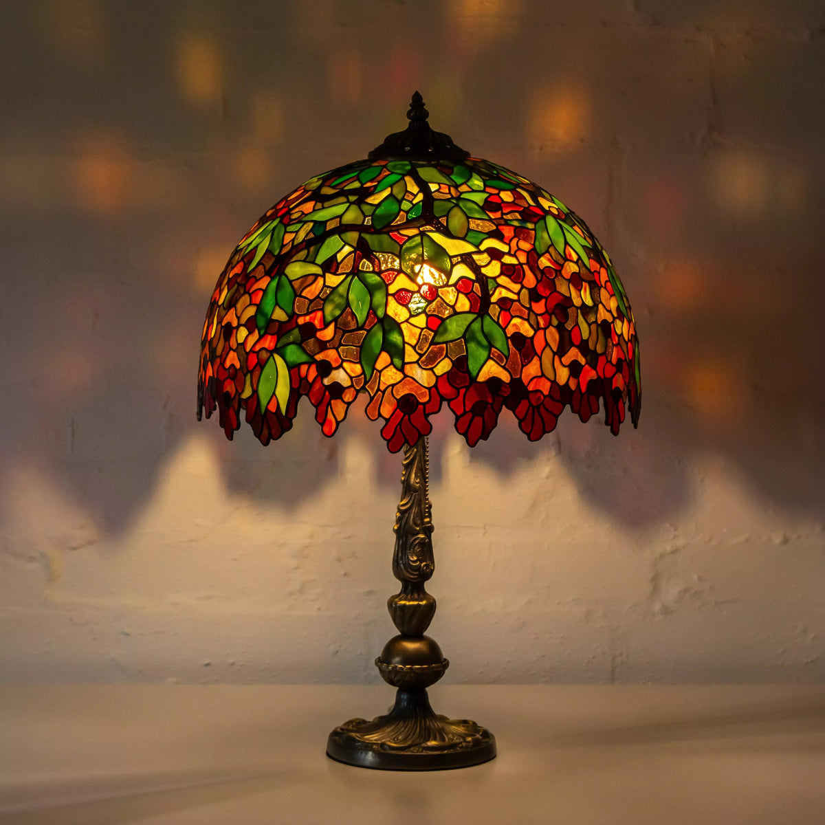 Full view of a stained glass table lamp with vibrant red flowers, green leaves, and bronze ornate base