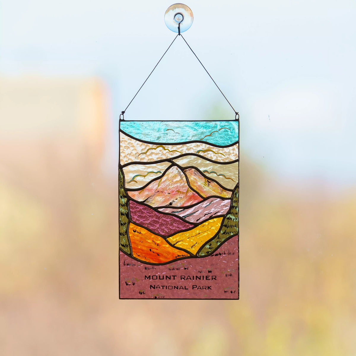A detailed Mount Rainier National Park stained glass panel hanging against a soft, sunlit outdoor background.