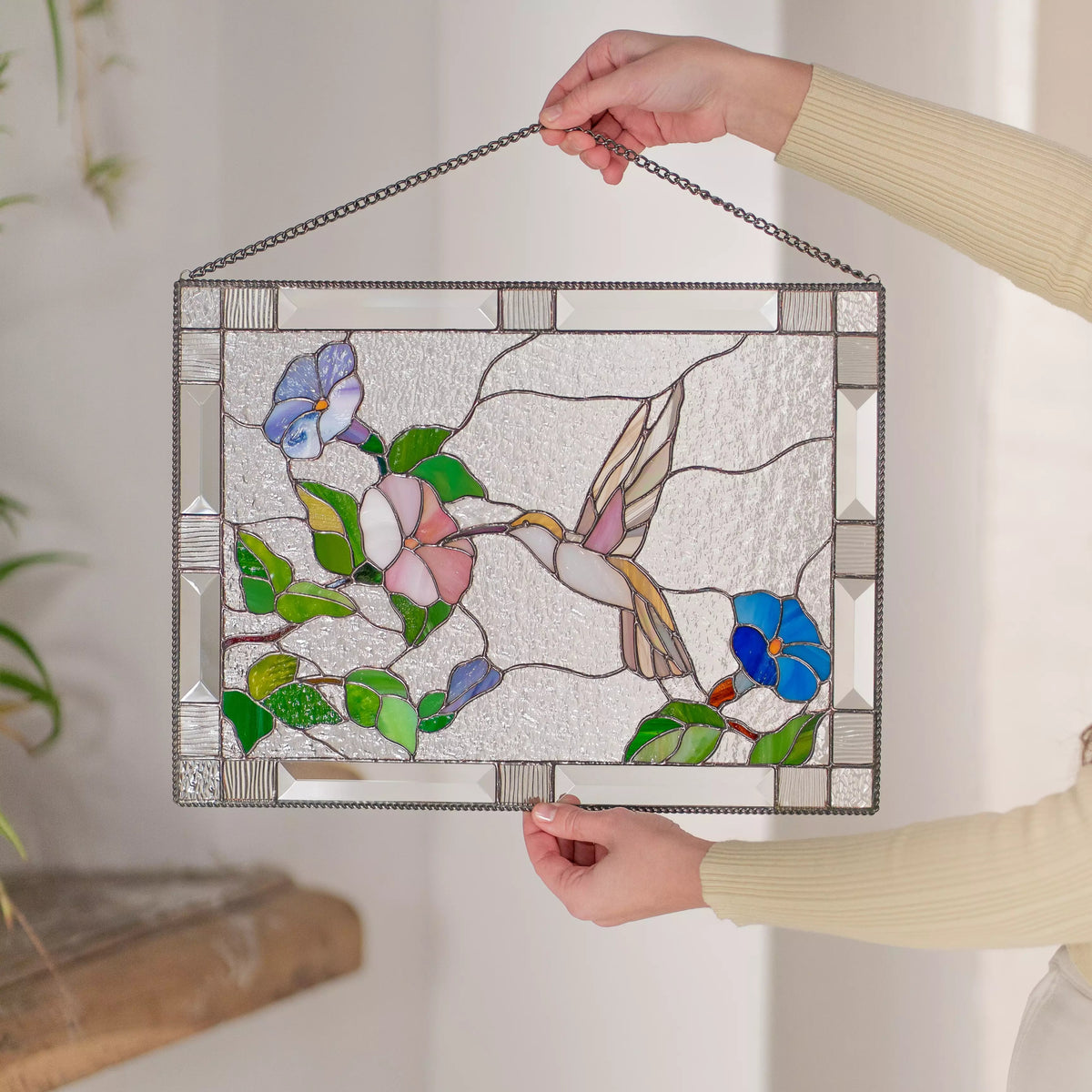 Stained glass panel featuring a hummingbird and flowers held by hands against a neutral background