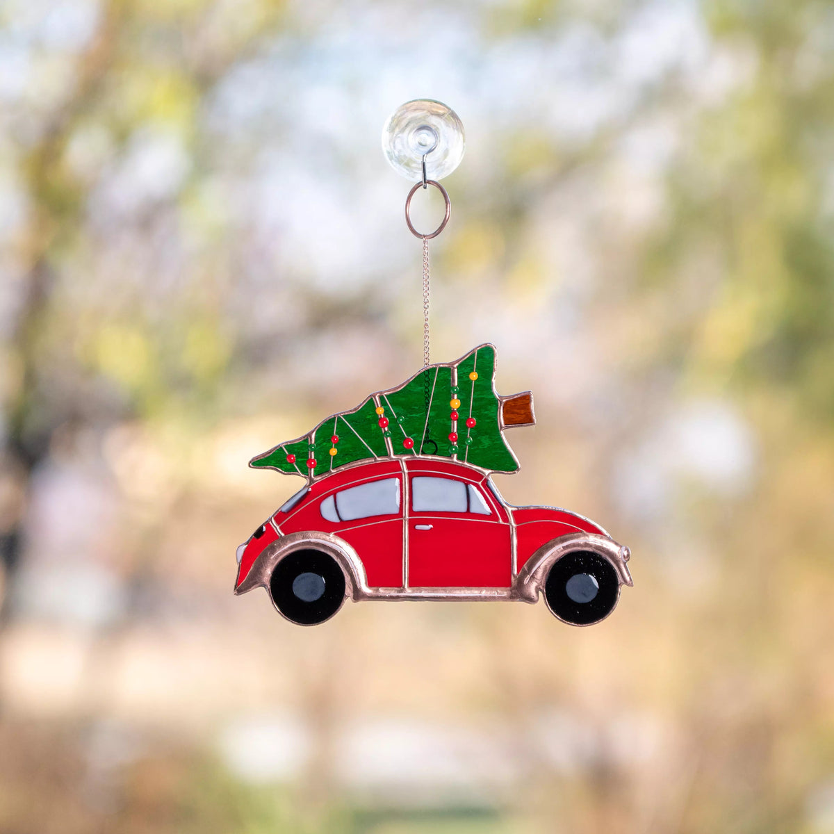 Stained glass window hanging featuring a red car with a Christmas tree on its roof, hanging against a blurred natural background.