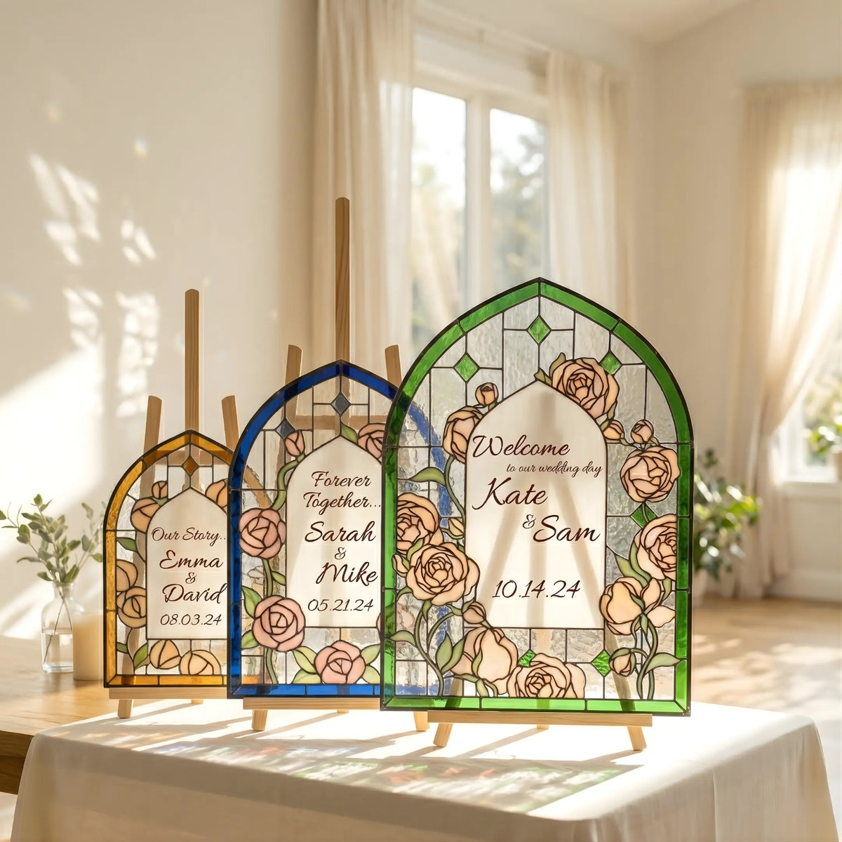 Three arched stained glass wedding sign panels with gold, blue, and green borders displayed on mini easels in a sunlit room.