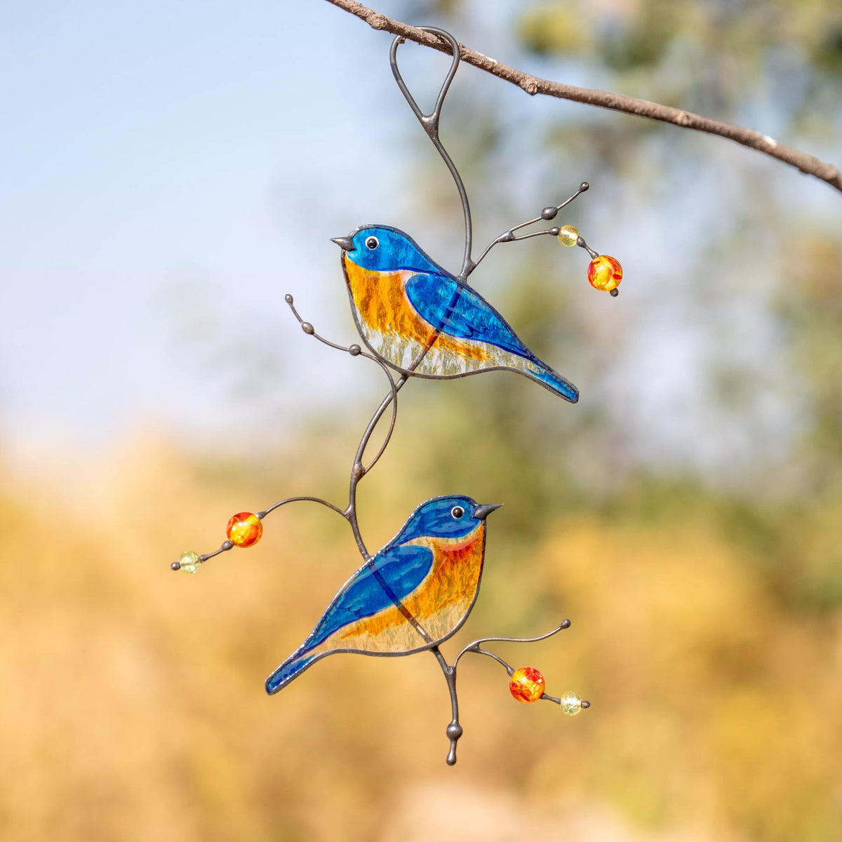 Two colorful stained glass bluebirds perched on a branch with a blurred natural background