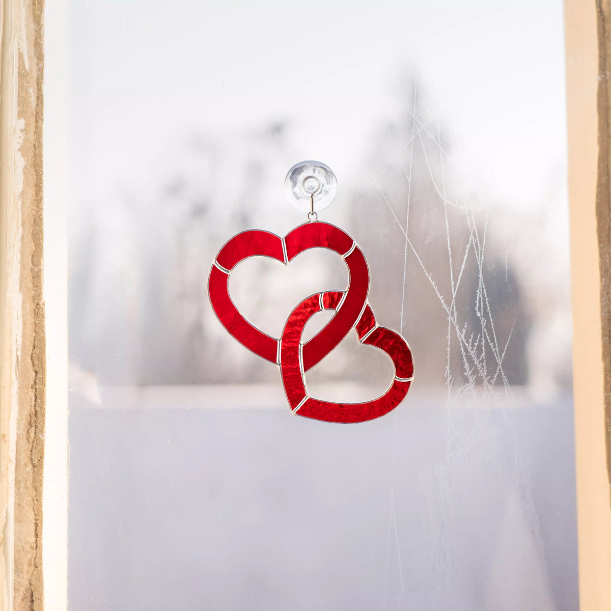 Red stained glass heart suncatcher attached to a window with a suction cup, glowing in natural daylight.