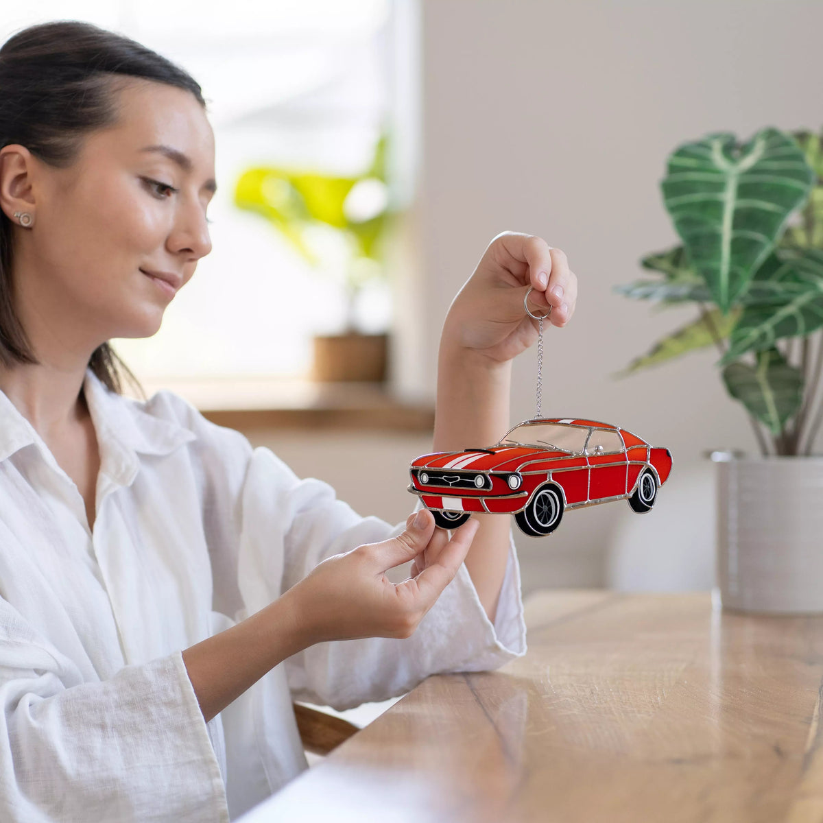 Woman holding a red car stained glass ornament in a home setting with plants in the background