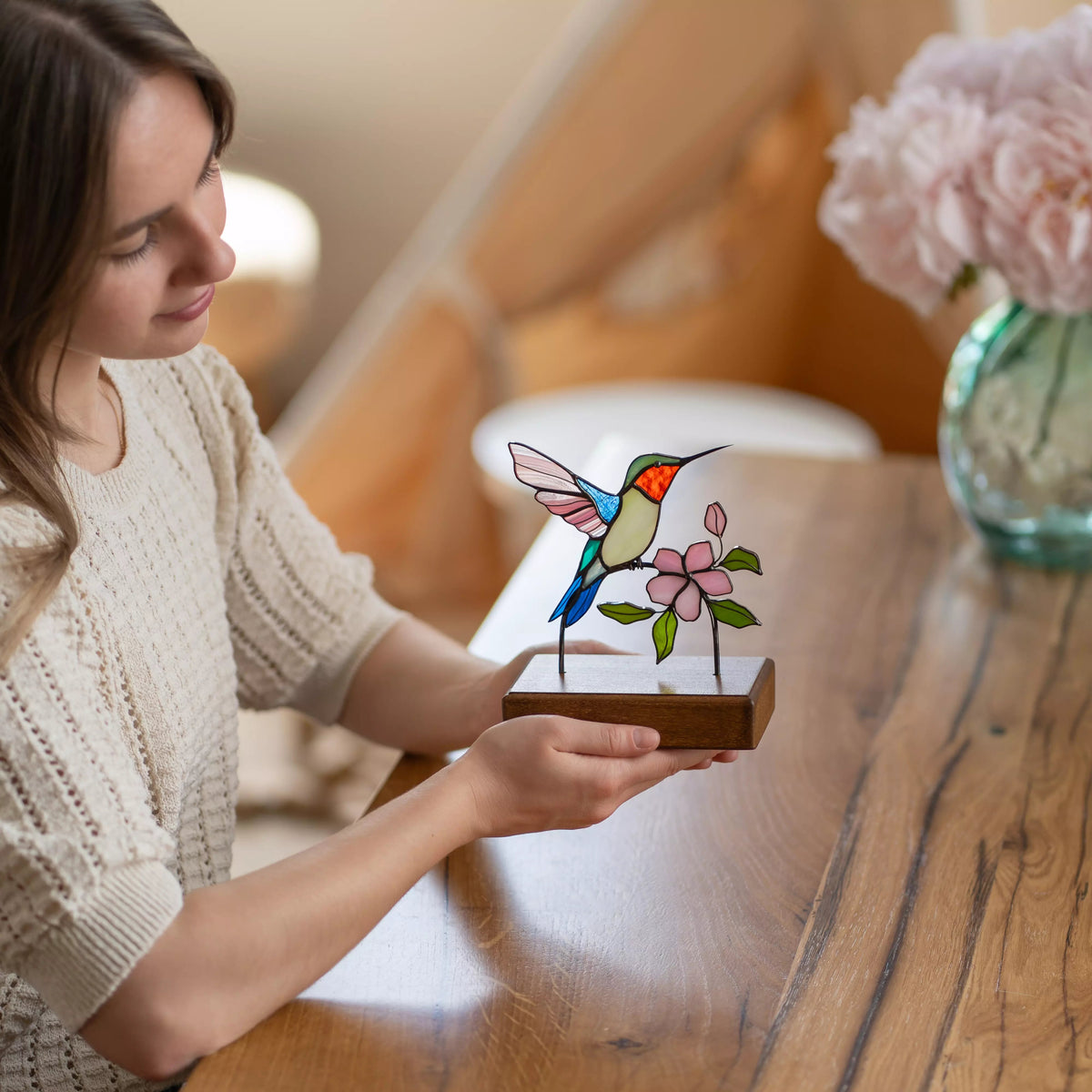 Woman holding a stained glass hummingbird and flower suncatcher on a wooden table.