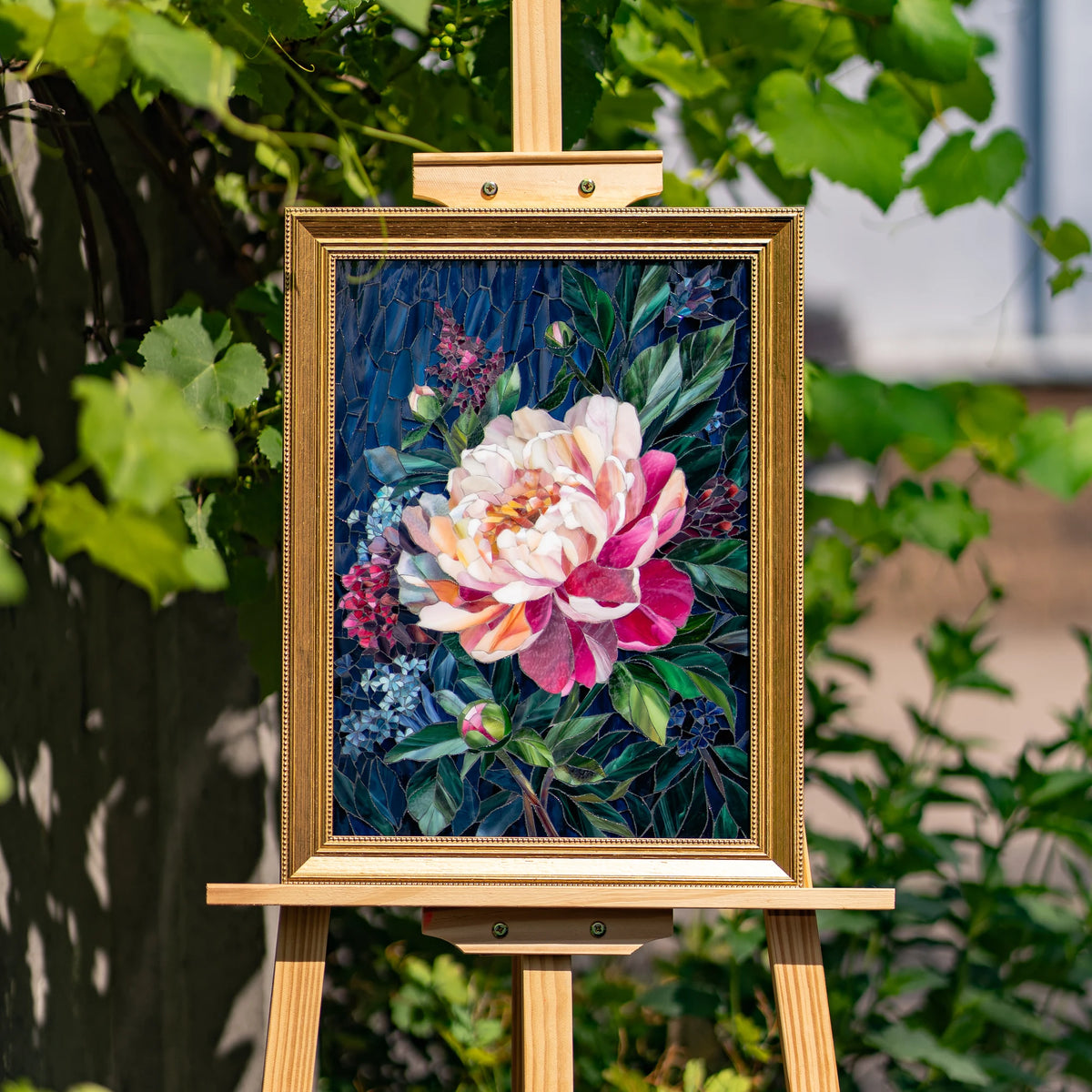 Gold-framed peony stained glass mosaic on a wooden easel in a sunlit garden, with rich pink petals and emerald glass leaves — handmade framed floral wall art that glows in daylight