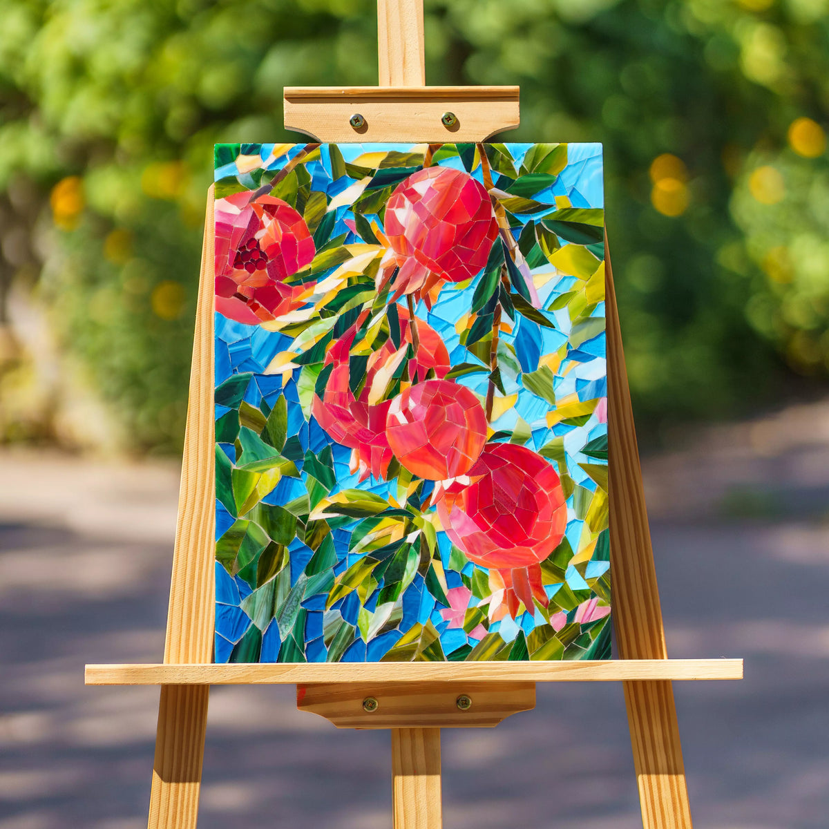 Pomegranate stained glass mosaic on a wooden easel outdoors, red glass fruit and green foliage against bright blue glass with sunlit greenery behind - handmade stained glass wall art and a thoughtful gift