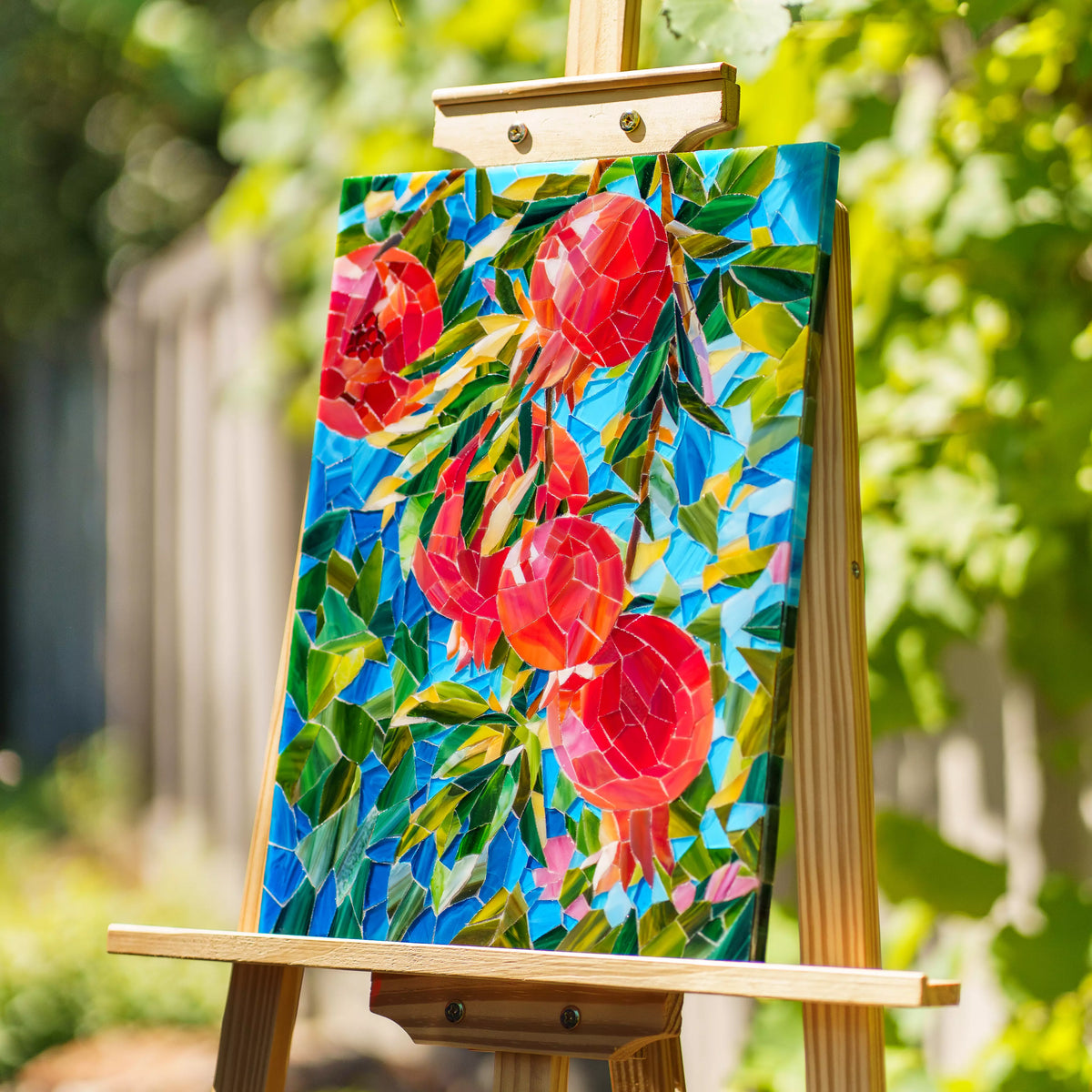 Handmade pomegranate stained glass mosaic on a wooden easel in a sunlit garden, red glass fruit and emerald leaves glowing against blue glass among the vines - vibrant stained glass wall art and a unique gift