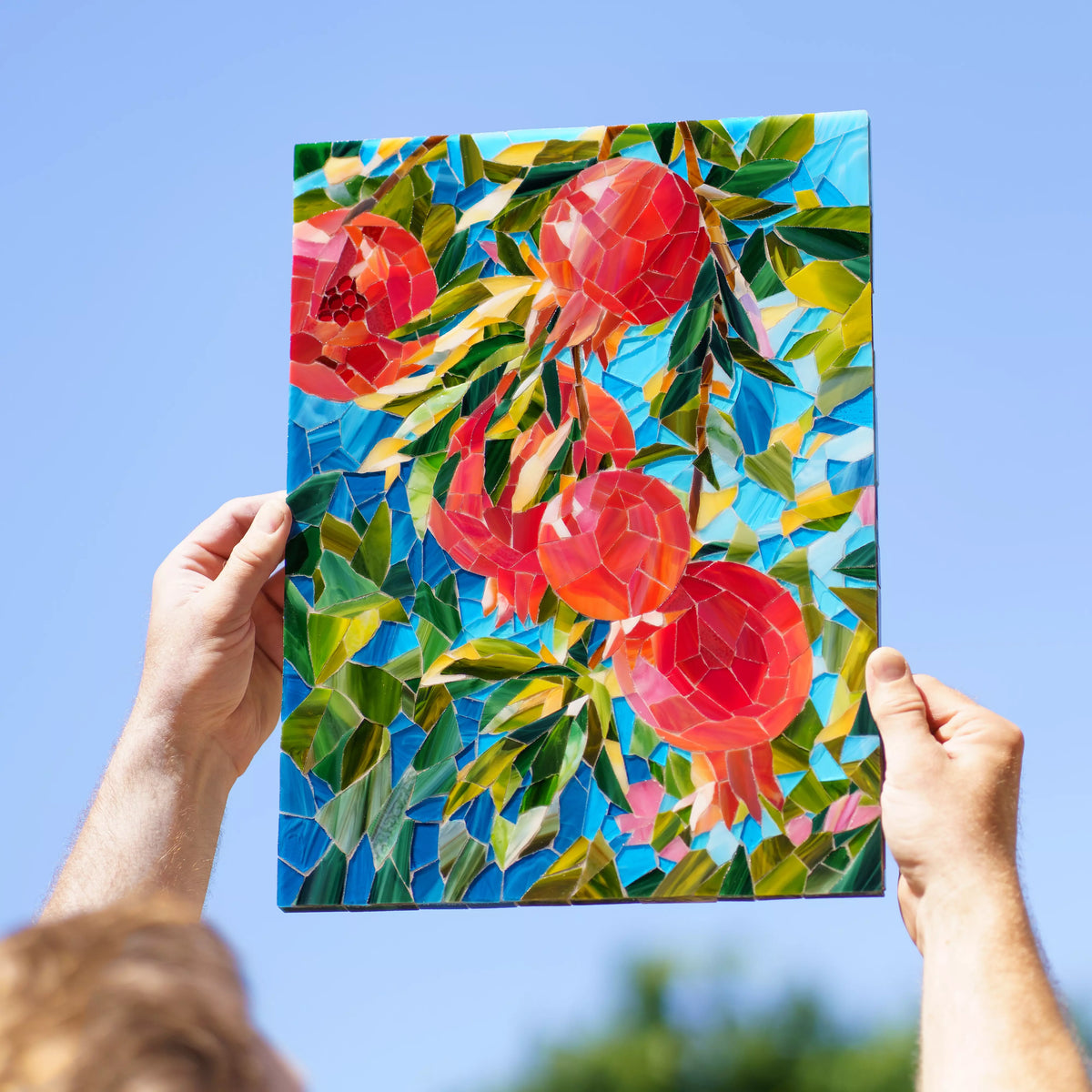 Hands holding the pomegranate stained glass mosaic up to a clear blue sky, sunlight glowing through the hand-cut red fruit and green leaves - handmade stained glass wall art that lights up in daylight
