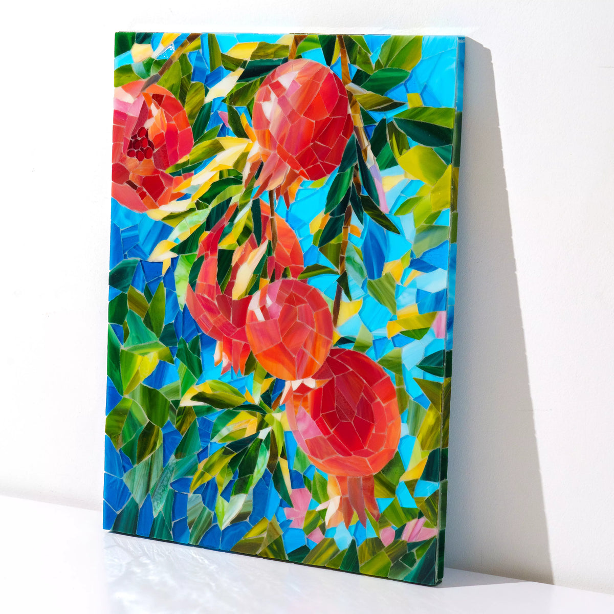 Angled view of the handmade pomegranate stained glass mosaic leaning on a white wall, revealing the glossy surface and depth of the hand-cut glass tiles - colorful stained glass wall art and statement piece