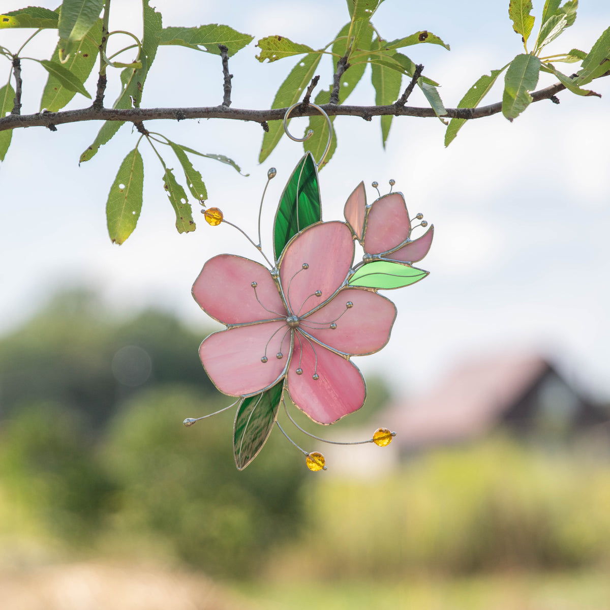 Stained glass sakura suncatcher with pink petals and green leaves hanging from a tree outdoors.