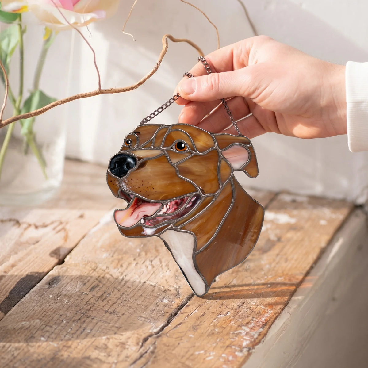 Hand holding a custom stained glass dog portrait by its chain above a rustic wooden windowsill, showing hand-painted eyes and nose details - a handmade pet suncatcher and heartfelt gift for pet lovers