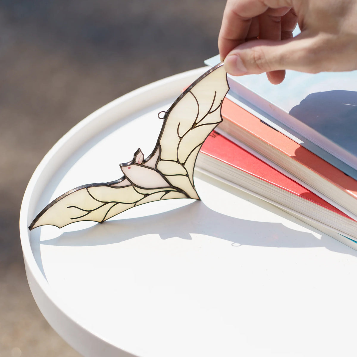 Hand lifting the white stained glass bat suncatcher from a round white side table stacked with colorful books in warm sunlight.