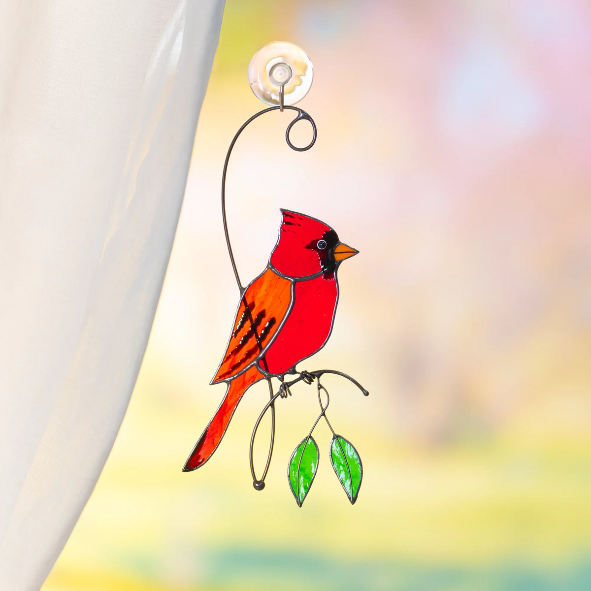 Red cardinal stained glass suncatcher on a suction cup by a sheer curtain, a brilliant red cardinal on a wire branch with green glass leaves against soft pastel light - handmade bird window decor