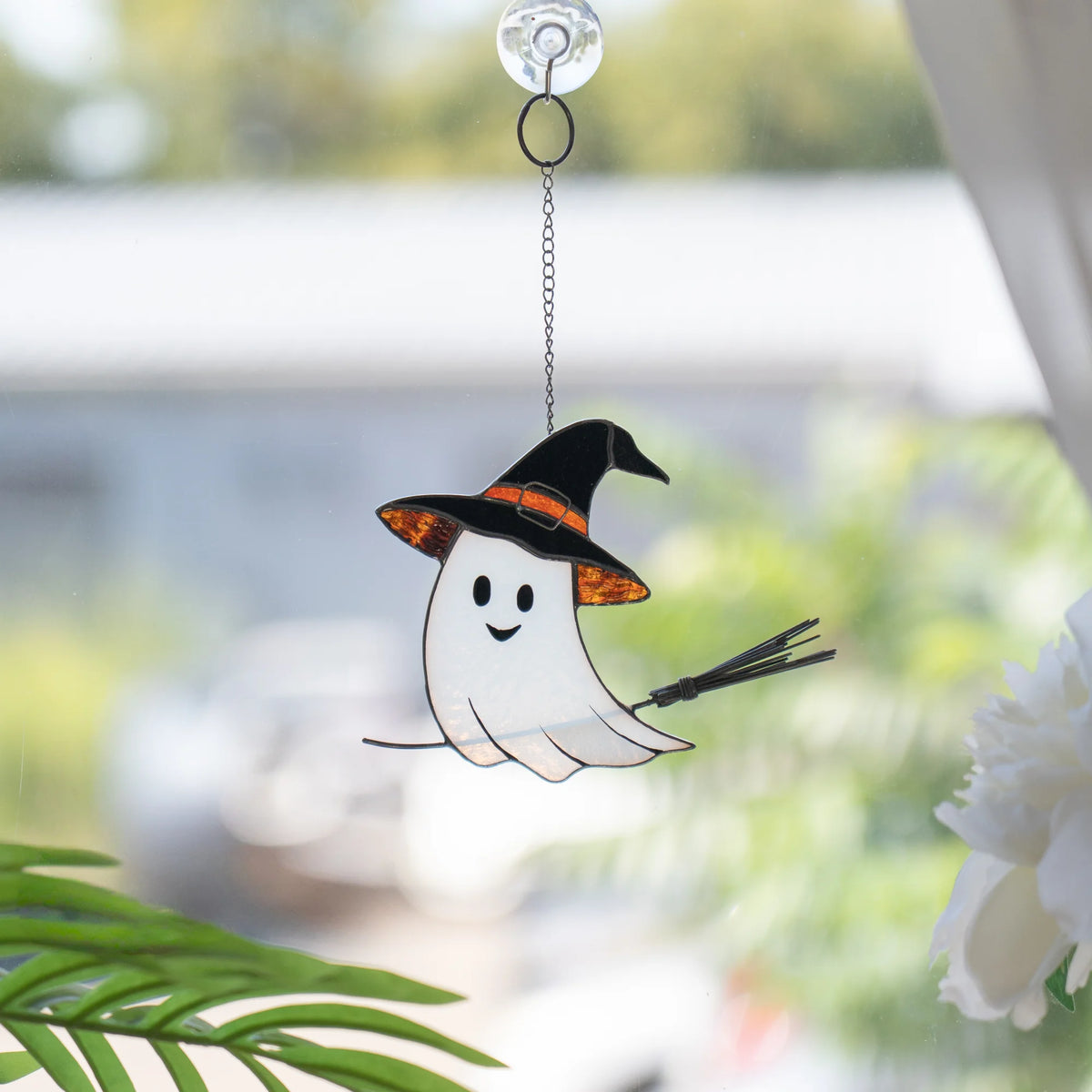 Stained glass ghost suncatcher hanging by a suction cup on a window, with green foliage and white flowers blurred behind - handmade Halloween glass art for the home