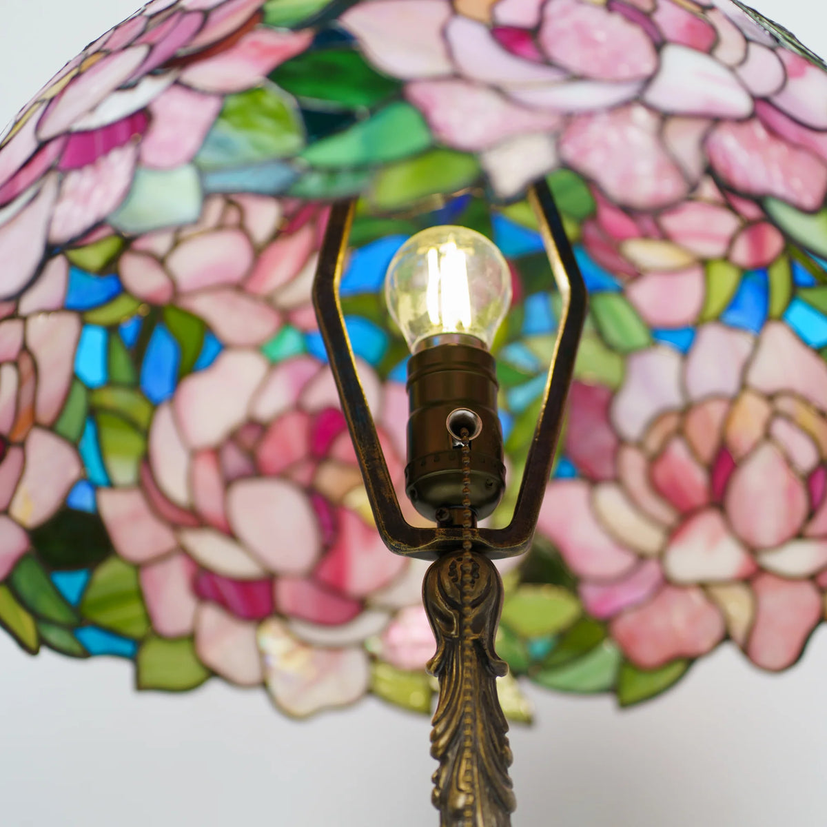 Close-up of a handmade stained glass peony lamp shade on white background, showing detailed pink and green glass petals, copper foil seams and a decorative brass finial on top